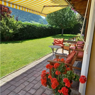 A cozy terrace with red geraniums and cushioned seating. The garden is green and a sunshade provides shade.