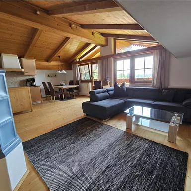 Ein gemütliches Wohnzimmer mit einem Holzdecken und großen Fenstern. Es gibt ein schwarzes Sofa, einen Glastisch und eine offene Küche.