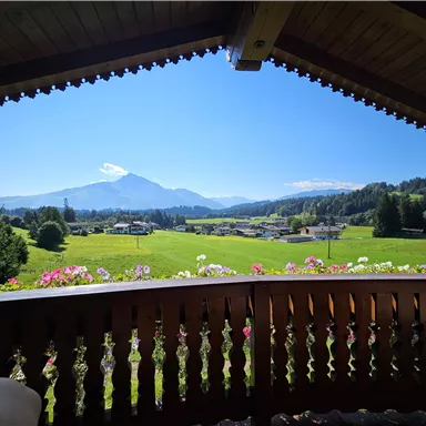 Eine malerische Berglandschaft mit grünen Wiesen und einem klaren blauen Himmel. Im Vordergrund ist ein Balkon mit Holzgitter und Blumen zu sehen.