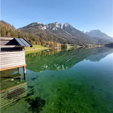 Ein klarer See umgeben von Bergen und Wäldern. Ein gemütliches Holzhaus steht am Ufer und spiegelt sich im Wasser.
