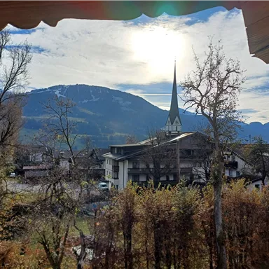 A picturesque view of mountains and a historic church tower. The sun shines through the clouds, giving the landscape a cozy atmosphere.