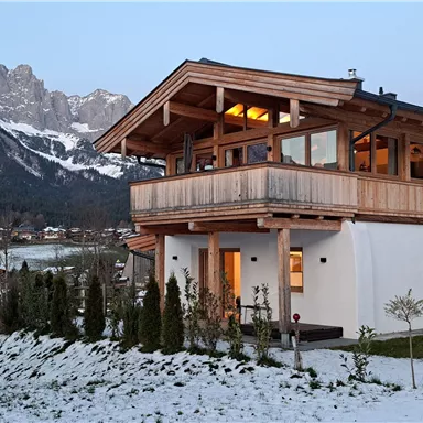 A modern house with wooden cladding and a balcony, surrounded by a snow-covered garden. Impressive mountains can be seen in the background.