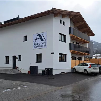 A modern building with the inscription "Apartment Alpenland". It is located in a quiet environment, surrounded by mountains.