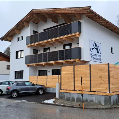 A modern building with several balconies and wooden cladding. In front of the building, some cars are parked.