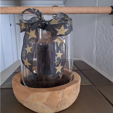 A decorative glass with a candle holder and a black-gold star ribbon, standing in a wooden bowl. The scene is warm and inviting.