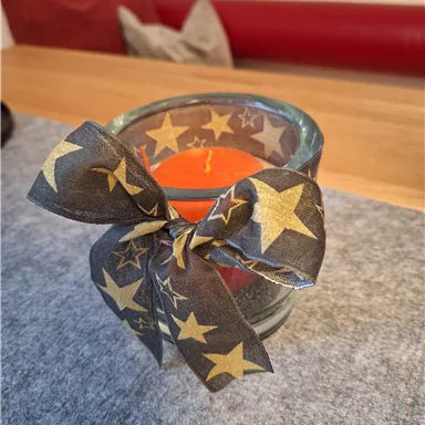 An orange candle in a glass container, surrounded by a black ribbon with golden stars. It is placed on a gray background.