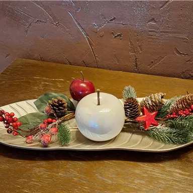 A festive decoration on a tray with a white candle, apples, pine cones, and green branches. The colors are warm and invite a cozy atmosphere.