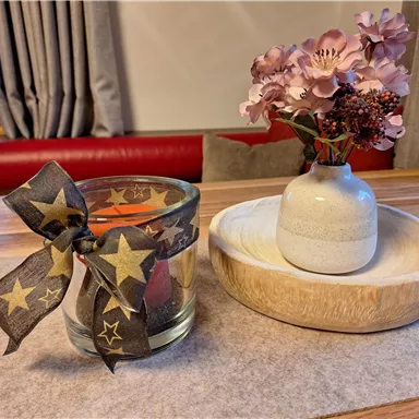 A decorative candle with a star ribbon and a small vase with flowers on a wooden plate. The background shows a cozy atmosphere.