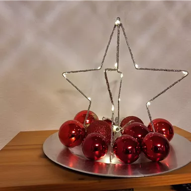 A decorative Christmas star made of wire with lights. Below are red Christmas ornaments on a round tray.