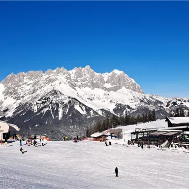 A snow-covered mountain landscape with clear blue sky. In the foreground, skiers and cabins can be seen.
