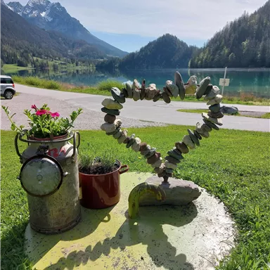 Ein Herz aus Steinen steht auf einer grünen Wiese neben einem blühenden Topf und einer dekorativen Milchkanne. Im Hintergrund sind Berge und ein klarer See zu sehen.