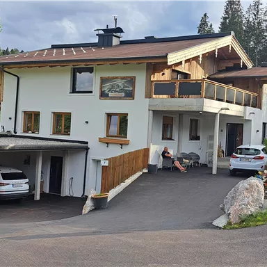 A modern house with several floors and a large balcony. In the foreground, parked cars and a green, well-kept environment can be seen.