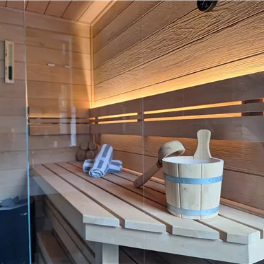 Eine moderne Sauna mit Holzverkleidung und sanfter Beleuchtung. Ein Holzbehälter mit einem Handtuch und einem kleinen Besen steht bereit.