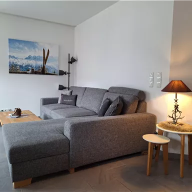Ein gemütliches Wohnzimmer mit einem grauen Sofa und einem kleinen Beistelltisch. An der Wand hängt ein Bild von Berglandschaft mit Skiern.
