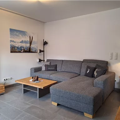 Ein modernes Wohnzimmer mit einem großen grauen Sofa und einem Holztisch. An der Wand hängt ein Landschaftsbild der Berge.