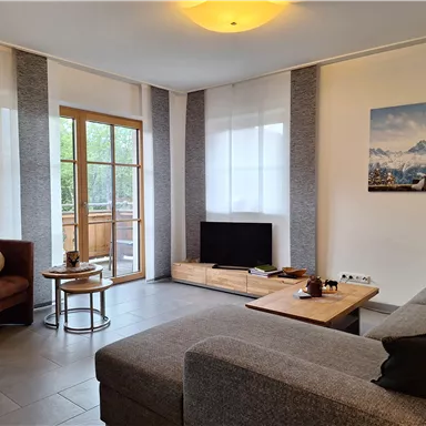 Ein modernes Wohnzimmer mit einer grauen Couch und einem kleinen Tisch. Große Fenster lassen viel Licht herein und bieten Zugang zu einem Balkon.