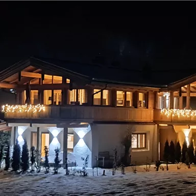 A cozy house at night, lit with festive lights. The snow in the foreground creates a wintry atmosphere.