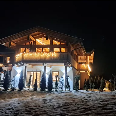 A nicely illuminated wooden house at night, surrounded by snow. The warm lights create a cozy atmosphere.