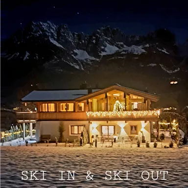 A cozy mountain cabin in the snow with festive lights. Impressive mountains can be seen in the background.
