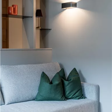 Ein modernes Wohnzimmer mit einem grauen Sofa und grünen Kissen. An der Wand sind Regale und eine elegante Wandlampe.