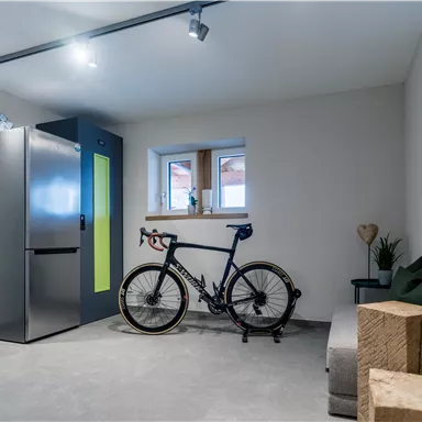 Ein moderner Raum mit einem Fahrrad, einem Kühlschrank und einem Fenster. Es gibt einige dekorative Pflanzen und eine gemütliche Sitzgelegenheit.