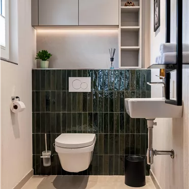 Ein modernes Badezimmer mit einer stilvollen grünen Fliese an der Wand. Es enthält ein WC, ein kleines Waschbecken und praktische Regale.