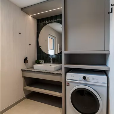 Ein modernes Badezimmer mit einer Waschmaschine und einem eleganten Waschbecken. Die Wände sind in hellen Farbtönen gehalten, und ein runder Spiegel reflektiert das Licht.