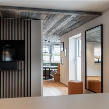 Ein modernes Wohnzimmer mit einem Fernseher an der Wand und einem großen Spiegel. Das Holzdecke und die Fenster schaffen eine helle, einladende Atmosphäre.