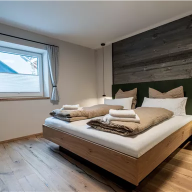 Ein modernes Schlafzimmer mit einem großen Doppelbett und Holzfußboden. Helle Vorhänge rahmen das Fenster ein und sorgen für eine gemütliche Atmosphäre.