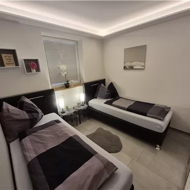 A modern bedroom with two single beds and elegant lighting. The walls are kept neutral, and there are decorative elements on the shelves.
