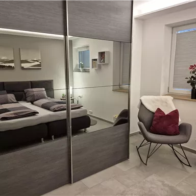 A modern bedroom with a large bed and a mirrored wardrobe. Next to the bed is a cozy armchair and a small side table.