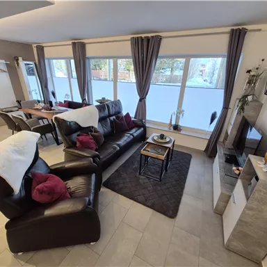 A modern living space with a comfortable sofa, a dining table, and large windows. The decor is warm and inviting, with decorative elements.