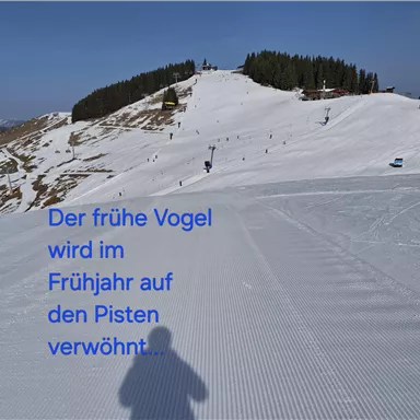 A snow-covered slope with many skiers in the background. The sun is shining on the snowy landscape.