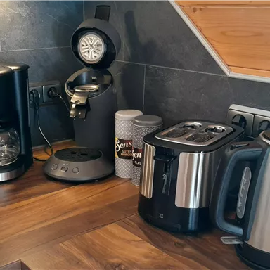 A modern kitchen corner with a coffee machine, a kettle, and a toaster. The wooden countertop gives the scene a cozy atmosphere.
