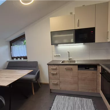 Eine moderne Küche mit Holzregalen und hellen Wänden. Der Raum hat ein großes Fenster und einen kleinen Essbereich mit Tisch und Stühlen.