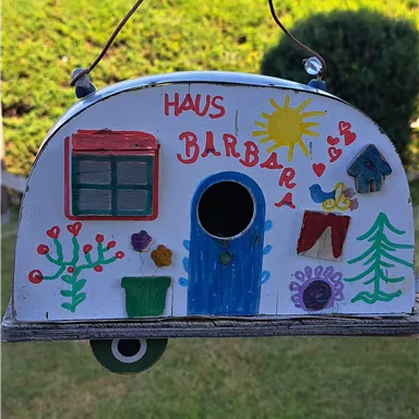 A colorfully decorated birdhouse in the shape of a caravan. It hangs in front of a green hedge and showcases a beautiful landscape in the background.