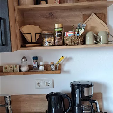A cozy kitchen corner with wooden shelves.
On top are jars, spices, and utensils, below are two coffee machines.