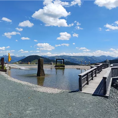 A quiet landscape with a wooden walkway over clear water. In the background are gentle hills and a blue sky with white clouds.