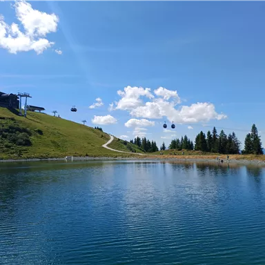 A clear blue lake with green meadows in the background. Cable cars travel across the landscape under a blue sky with few clouds.