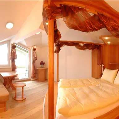 Ein gemütliches Zimmer mit einem Holzbett und hellen Laken. Zweisitzen und ein Tisch bieten eine angenehme Atmosphäre.