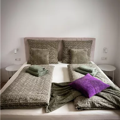 A modern bed with two pillows and various blankets. On the bed are green towels and a purple pillow.