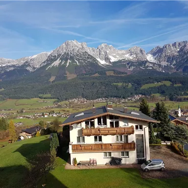 A picturesque mountain landscape with snow-covered peaks and lush green meadows. In the foreground stands a modern house with balconies.