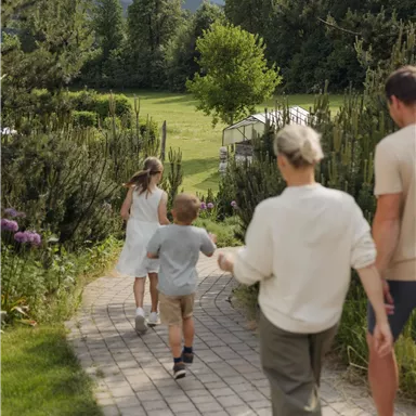 A family walks along a paved path, surrounded by green trees and flowers. In the background, gentle mountains can be seen.