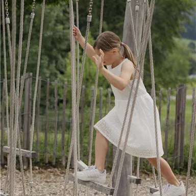 A girl in a white dress is playing on a playground. She is crossing a wobbly rope bridge.