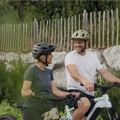 A couple sits on bicycles and smiles at each other. In the background, green bushes and a wooden fence can be seen.