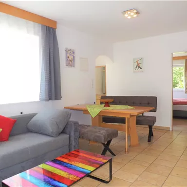 Ein modernes Wohnzimmer mit einer gemütlichen Couch, einem Esstisch und buntem Dekor. Das Zimmer hat große Fenster und einen hellen, einladenden Stil.