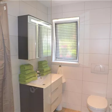 Ein modernes Badezimmer mit hellen Fliesen und einem Fenster. Handtücher in frischem Grün liegen ordentlich gestapelt.