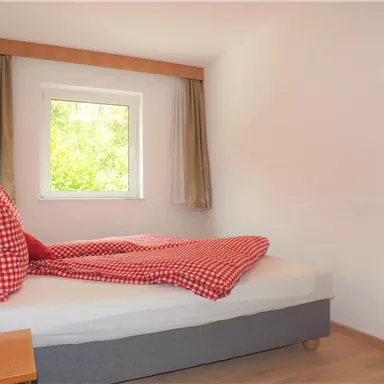 Ein gemütliches Schlafzimmer mit einem Bett und einem Fenster. Die Bettwäsche hat ein rot-weißes Karomuster und es gibt einen Nachttisch daneben.