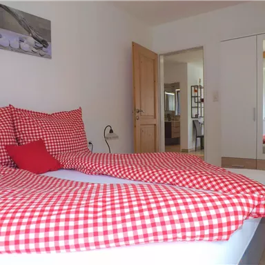 Ein gemütliches Schlafzimmer mit einem großen Bett, das mit karierter Bettwäsche in Rot und Weiß dekoriert ist. Im Hintergrund sieht man einen Schrank und Fenster mit Blick ins Grüne.