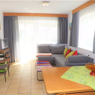 Ein heller Wohnzimmerbereich mit einer grauen Couch und bunten Kissen. Ein Tisch mit einer grünen Tischdecke und Fenster, die viel Tageslicht hereinlassen.